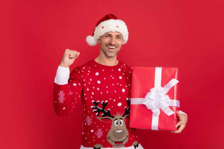 Christmas gift, present box. Studio portrait of guy wears santa hat and winter sweater. Man with christmas hat over isolated red background.の写真素材