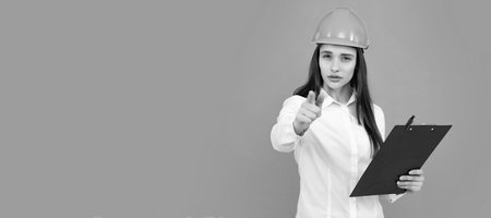 Serious woman in protective helmet with clipboard isolated on grey background. Young woman construction manager. Woman, female worker in hardhat helmet pointing to you.の写真素材