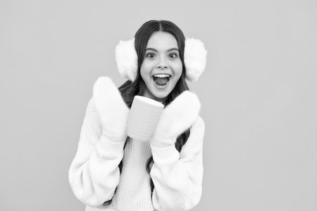 Excited girl in winter warm earmuff ear-flaps and sweater with take away cup of cappuccino coffee or tea. Child with plastic takeaway mug, morning drink cocoa beverage.の写真素材
