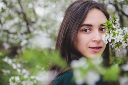 Young caucasian girl with dark hair in white t-shirt and green coat near beautiful white cherry blossom tree spring nature backgroundの写真素材
