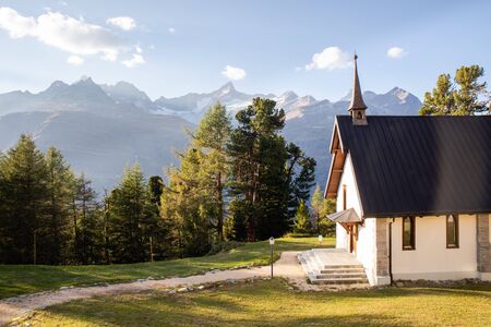 White house with dark wooden roof near forest with beautiful Swiss Apls mountains in background