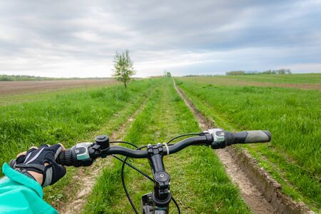 Hand in cycling gloves holding steering wheel of bicycle riding summer road in green field, active...