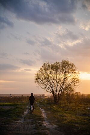 Girl in jacket and cap riding bicycle in rural field road into susnetの写真素材