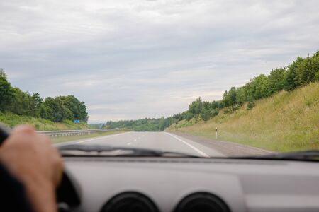 View on long road with fields and trees from front car window with stiring wheel, road trip travel...