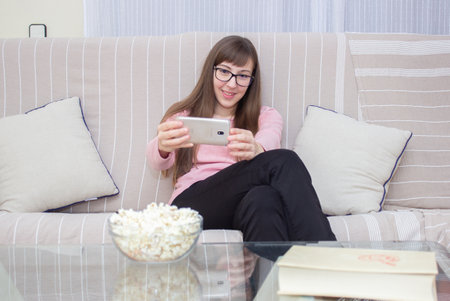 woman sitting on the sofa making video calls with her mobile and a bowl popcorn on the tableの写真素材