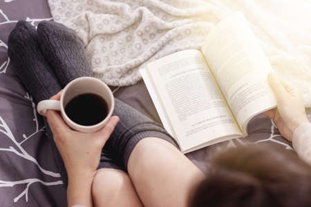 a woman drinking a black coffee and reading a book on the bed with a blanket.の写真素材
