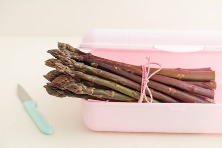 Bunch of fresh purple asparagus in pink plastic container with kitchen knifeの写真素材