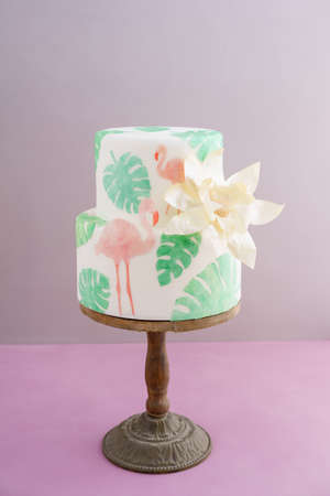 Two tiered tropical wedding cake with fondant, tropical wafer paper leaves and flamingos with a big golden statement flower on wooden cake standの写真素材
