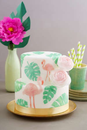 Two tiered tropical wedding cake with fondant, tropical wafer paper leaves and flamingos with ranunculus flowers on golden cale platterの写真素材
