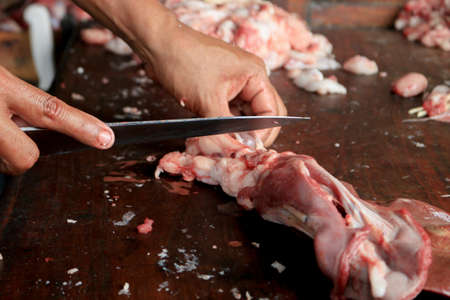 Butcher cutting raw goat leg meat on a wooden chopping Board with knife.