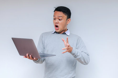 Wow face of Young Asian man shocked what he see in the laptop when working isolated grey background wearing grey shirt.の写真素材
