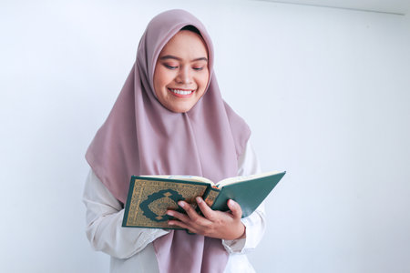 Young Asian Islam woman wearing headscarf is praying or read Quran - the holy book of Islam with smile and serious face. Indonesian woman on gray background.の写真素材