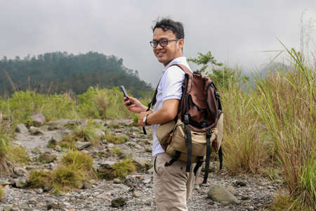 Young asian backpacker man is happy and smile when he looking smartphone when traveling in mountain
