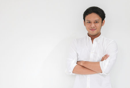 Young Asian man wearing white shirt is holding hands crossed with happy smile and confidence. Successful Indonesian man on gray background.の写真素材