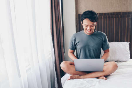 Happy Asian man is working with his laptop on his bed. Concept of freelancer successful lifestyle.の写真素材
