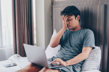 Tired stressed young Asian man feeling sleepy and tired while using laptop on the bed in bedroom. Hard work concept.の写真素材