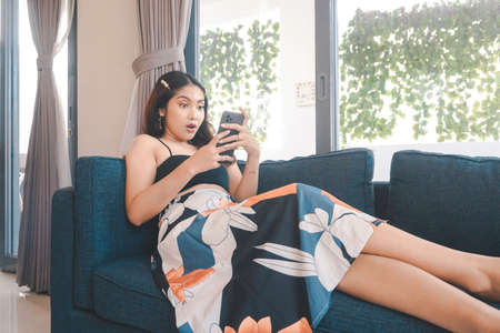 Young Asian woman spend her free time at home sitting on a cozy sofa in the living room surprised while looking on her smartphone.の写真素材
