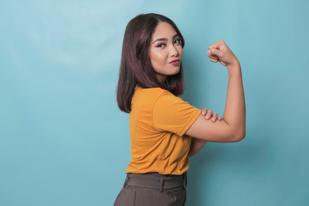 Young beautiful girl wearing casual yellow t-shirt standing over isolated blue background showing arms muscles smiling proud. Fitness concept.の写真素材