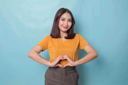 Attractive young Asian woman feels happy and romantic shapes heart gesture expresses tender feelings wears casual yellow t-shirt against blue background. People affection and care conceptの写真素材