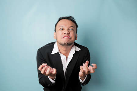 Angry and mad face of Asian businessman in black suit on isolated blue background.