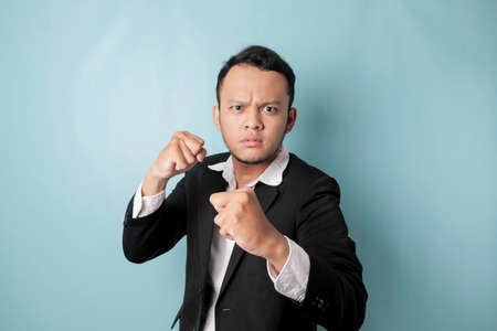 A portrait of a young Asian businessman looks angry and holding his fist ready to punch, isolated by blue backgroundの写真素材