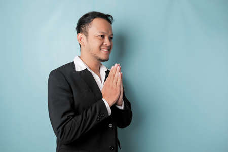 Smiling young Asian businessman wearing black suit gesturing greeting or namaste isolated over blue backgroundの写真素材