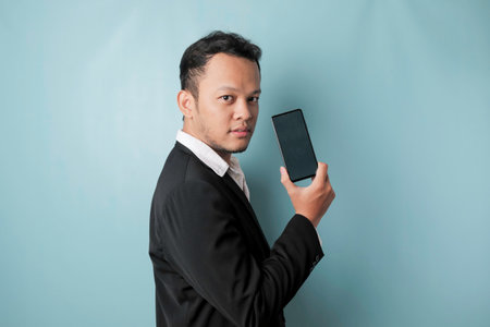 A young Asian businessman in a black suit feels serious and focus holding a smartphone and showing the copy space on its screen in hand.の写真素材