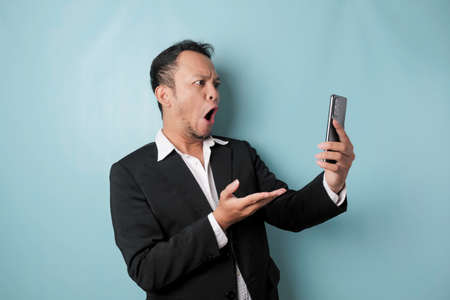 Surprised Asian businessman wearing black suit holding his smartphone, isolated by blue background