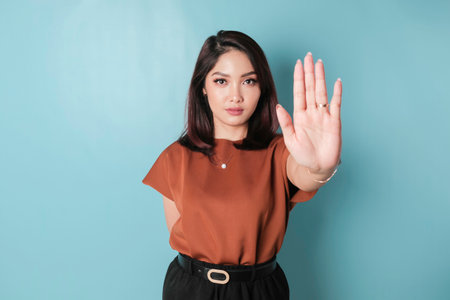 Young Asian woman wearing brown shirt over blue isolated background doing stop sign with palm of the hand.の写真素材