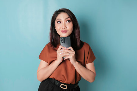 Portrait of a thoughtful young woman wearing brown shirt looking aside while holding smartphone isolated over blue backgroundの写真素材