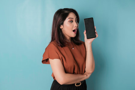 Surprised woman wearing a brown shirt showing copy space on her smartphone, isolated by a blue backgroundの写真素材