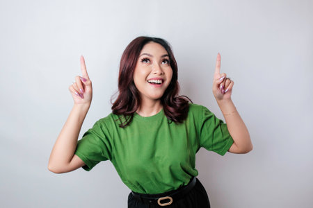 Excited Asian woman wearing green t-shirt pointing at the copy space upside her, isolated by white backgroundの写真素材