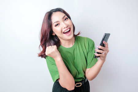 A young Asian woman with a happy successful expression wearing green t-shirt and holding smartphone isolated by white backgroundの写真素材