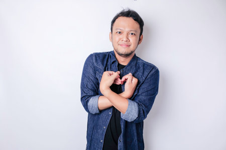 An attractive young Asian man wearing a navy blue shirt feels happy and a romantic shapes heart gesture expresses tender feelingsの写真素材