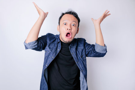 Shocked Asian man wearing blue navy shirt pointing at the copy space on top of him, isolated by white backgroundの写真素材