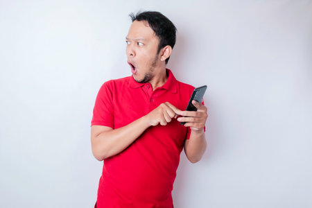 Surprised Asian man wearing red t-shirt pointing at his smartphone, isolated by white backgroundの写真素材