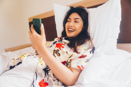Happy young woman checking on her phone while lying in bedの写真素材