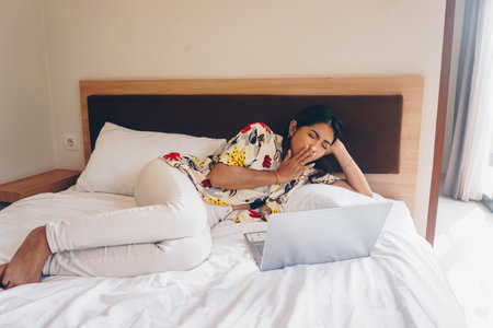 A sleepy young woman yawning while working from the bed.