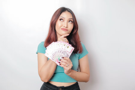 A thoughtful young woman is wearing blue t-shirt and holding cash money in Indonesian rupiah isolated by white backgroundの写真素材