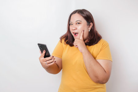 Surprised Asian big size woman wearing yellow t-shirt pointing at her smartphone, isolated by white...
