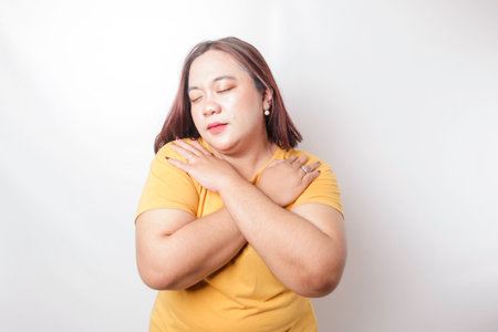 Young beautiful Asian big sized woman wearing a yellow t-shirt over white background hugging herself happy and positive, smiling confident. Self-love and self-careの写真素材
