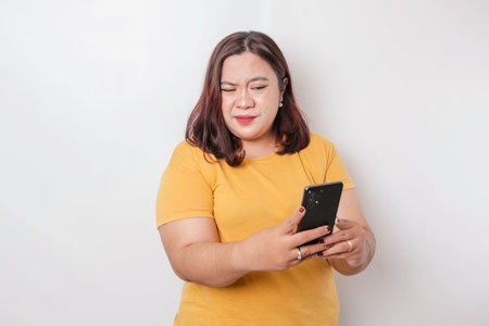 The angry and mad face of big sized Asian woman in yellow shirt while holding her phone on isolated white background.の写真素材