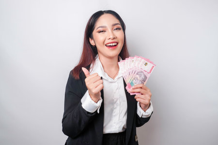 Excited Asian businesswoman wearing black suit gives thumbs up hand gesture of approval and holding money in Indonesian Rupiah, isolated by white backgroundの写真素材