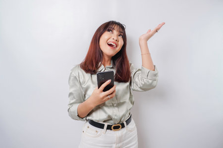 Excited Asian woman wearing sage green suit pointing at the copy space on top of her while holding her phone, isolated by white backgroundの写真素材