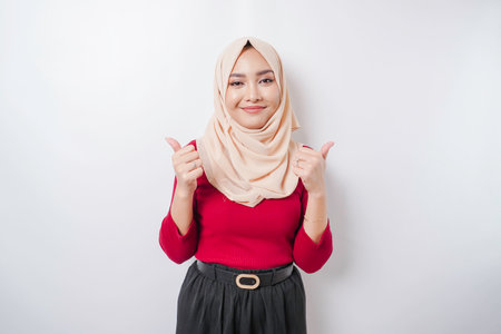 Excited Asian woman wearing a hijab gives thumbs up hand gesture of approval, isolated by white backgroundの写真素材