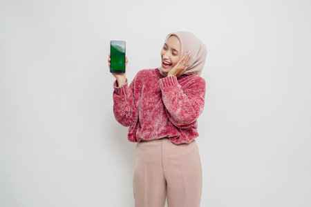 Excited Asian Muslim woman wearing pink sweater and hijab pointing at the copy space beside her...