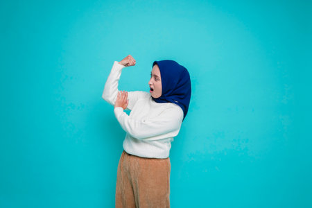 Excited Asian Muslim woman wearing a white shirt and hijab showing strong gesture by lifting her...