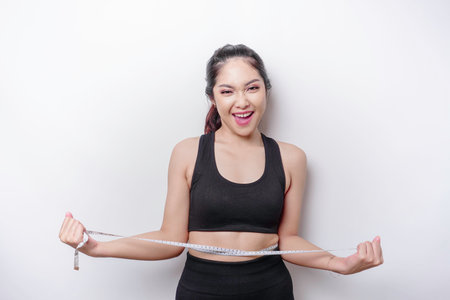 Portrait of cheerful young Asian woman in sportswear measuring waist with tape, feeling happy about...