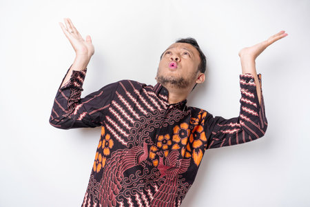 Shocked Asian man wearing batik shirt pointing at the copy space on top of him, isolated by white...