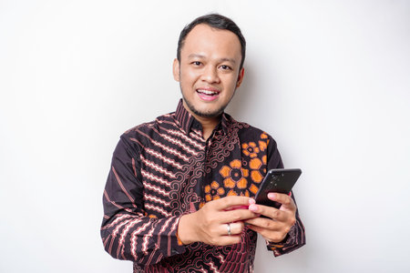 A portrait of a happy Asian man wearing batik shirt and holding his phone, isolated by white...